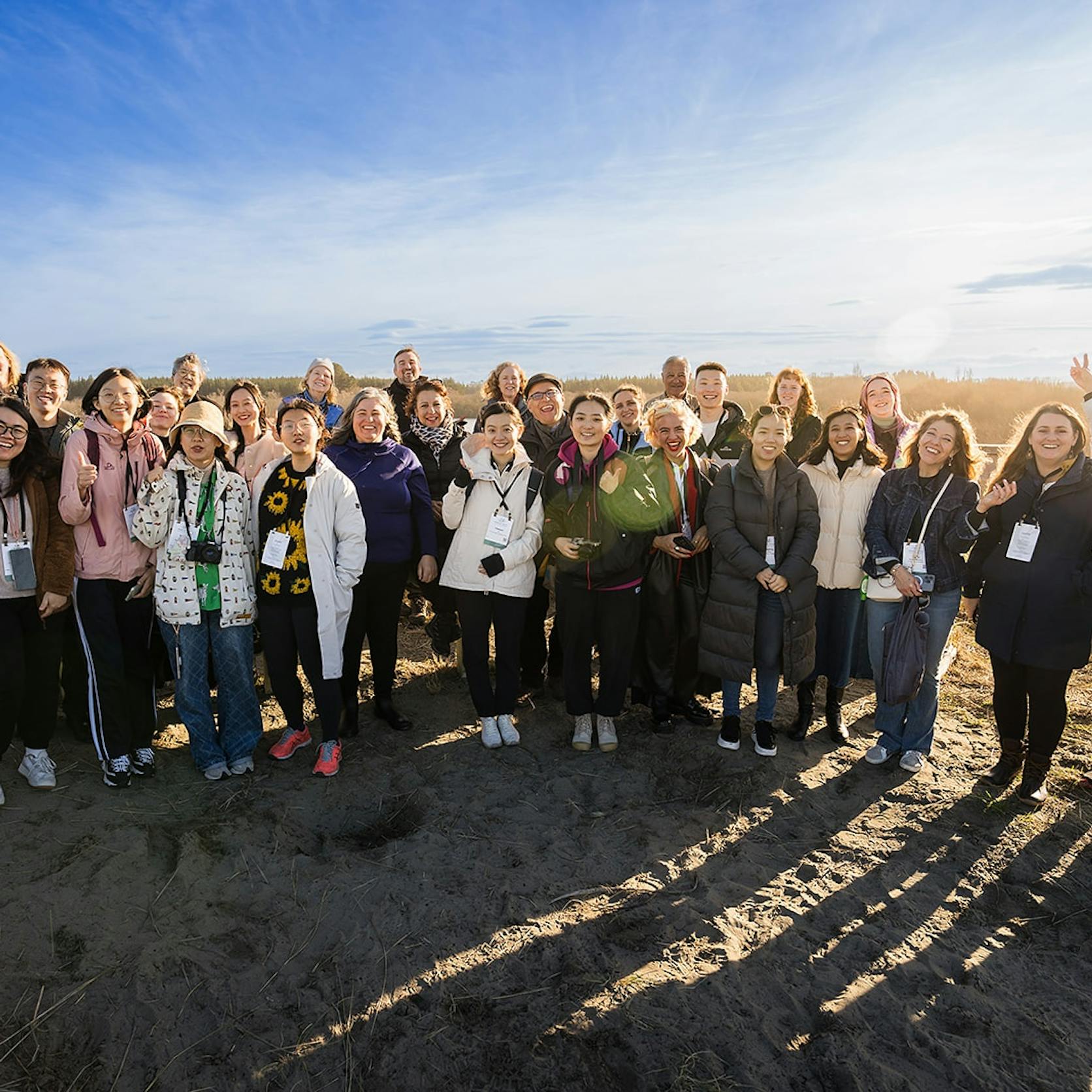 A diverse group of people stands together outdoors, smiling and interacting, showcasing the welcoming spirit of Te Pae. The sun sets behind them, illuminating the sandy environment and surrounding greenery.